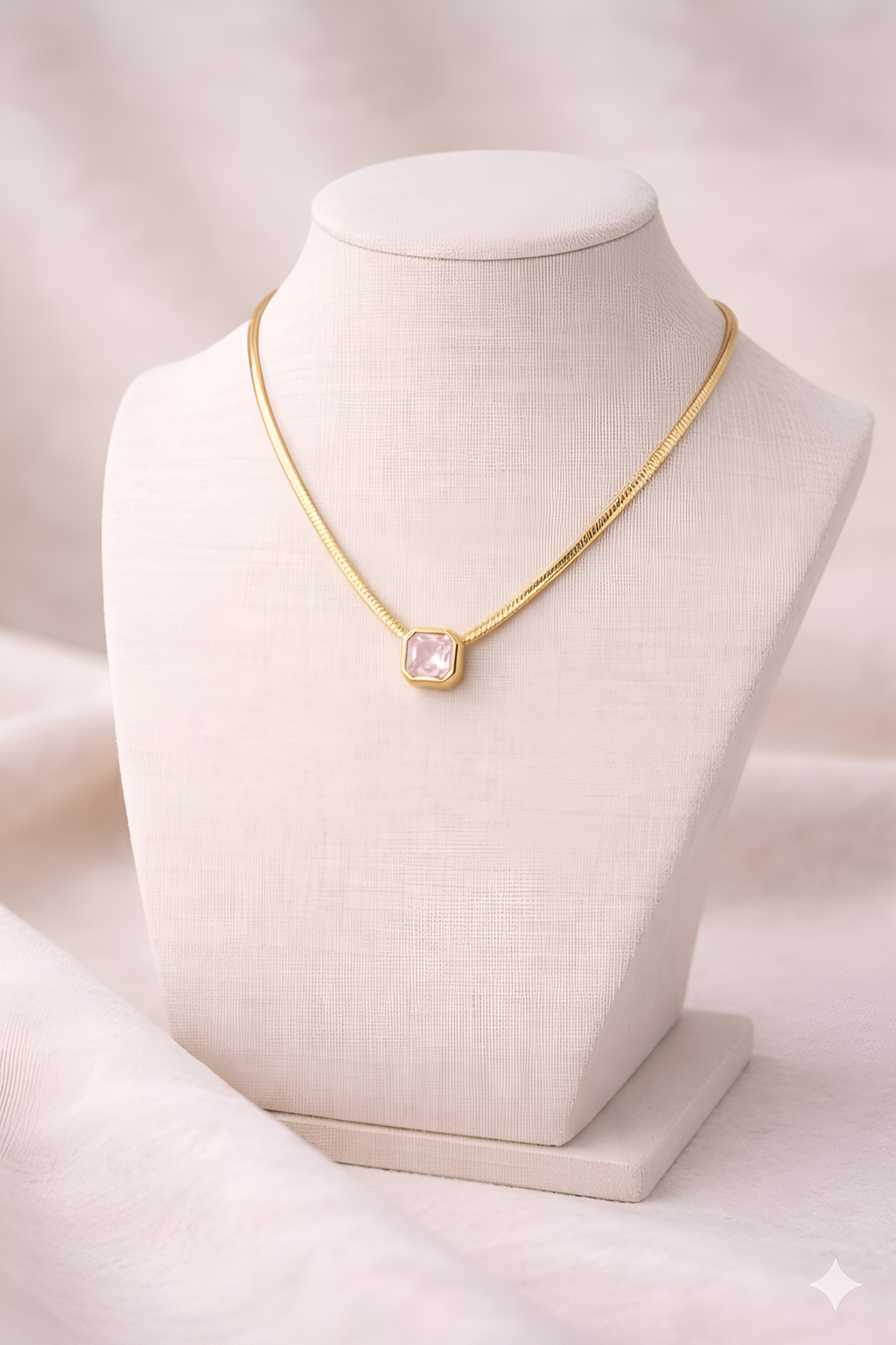 Gold snake chain necklace with blush pink rectangular pendant on ivory stand