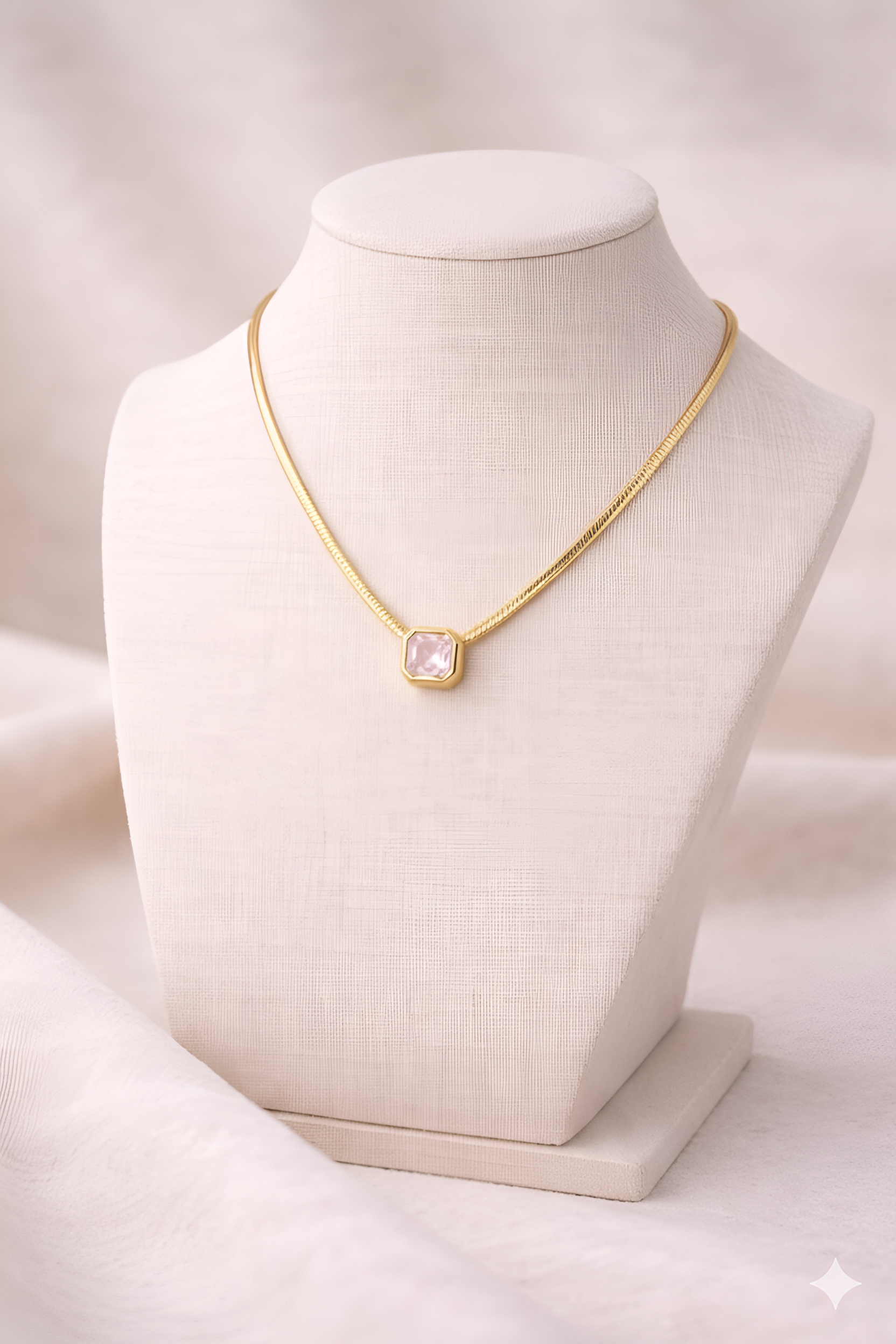 Gold snake chain necklace with blush pink rectangular pendant on ivory stand