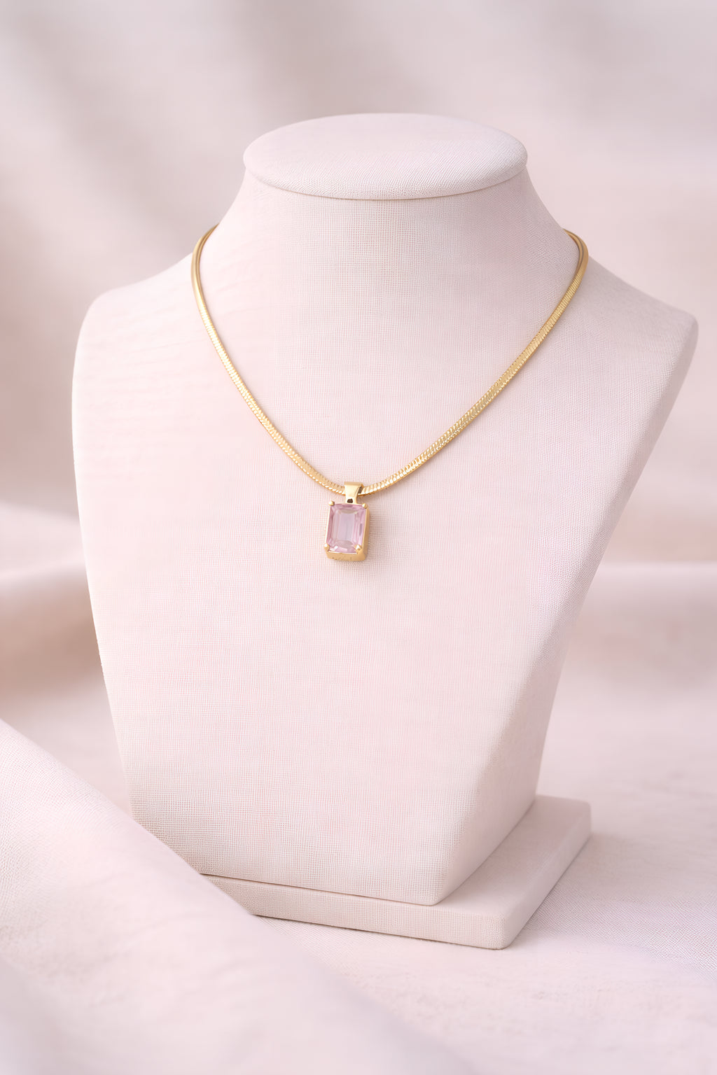 Gold snake chain necklace with blush pink rectangular crystal pendant on ivory stand